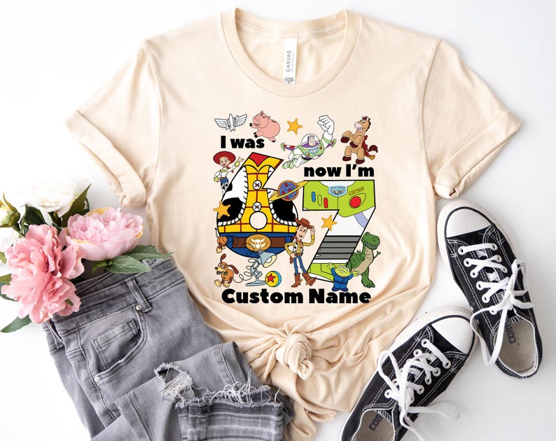 May include: A beige t-shirt featuring a colorful graphic with the text "I was now I'm" and the number 7. The graphic includes cartoon characters and the words "Custom Name". Black canvas sneakers and gray jeans are also in the image.