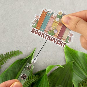 May include: A clear badge reel with a sticker featuring a stack of books and flowers. The sticker reads "BOOKTROVERT" in brown letters. The books are in shades of orange, teal, pink, and yellow. The badge reel is attached to a clear plastic clip.