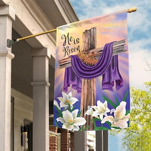 May include: A decorative Easter flag featuring a wooden cross draped with a purple cloth and a crown of thorns. The flag has the text "He is Risen" and is adorned with white lilies against a colorful sky background.