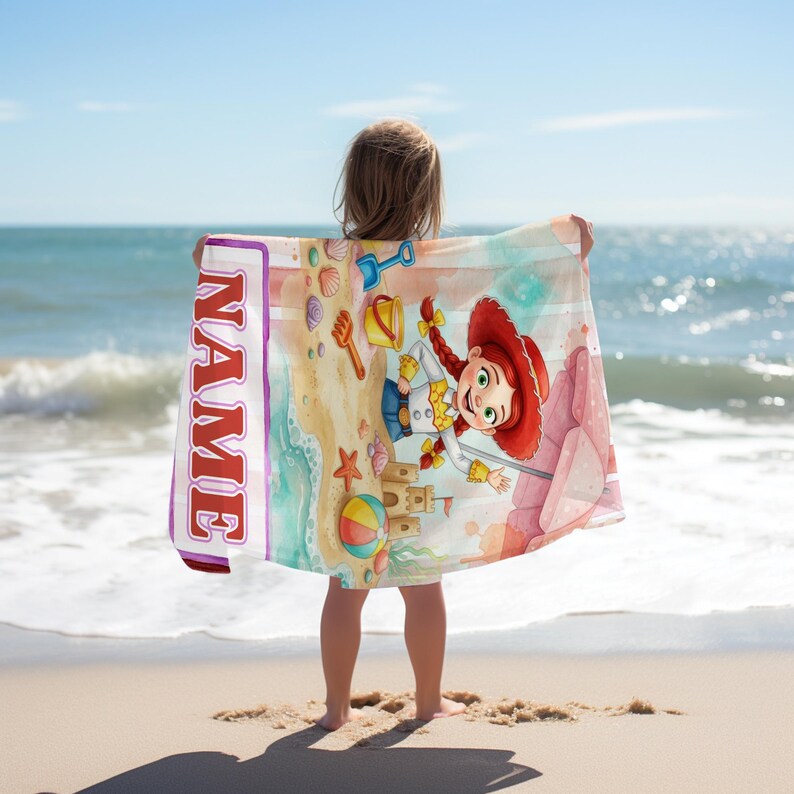 May include: A rectangular beach towel featuring a cartoon cowgirl character with red braided hair, a sandcastle, a beach ball, and a shovel on a sandy beach. The towel includes a vertical white stripe on the left side with the red text NAME.