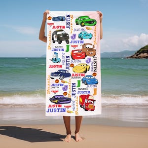 May include: A white beach towel with cartoon car illustrations and the name "Justin" printed on it. The towel has a light pink background and is held up on a beach with the sea in the background.