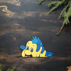 May include: A yellow and blue cartoon fish with green eyes and a smiling expression. The fish has blue stripes and fins. The image is set against a dark wood background with green foliage.