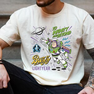 May include: A cream-colored t-shirt featuring a graphic of Buzz Lightyear in a flying pose. The design includes the text "TO INFINITY AND BEYOND" in green, "Buzz" in yellow script, and "LIGHTYEAR" in purple, along with a planet and star icons.