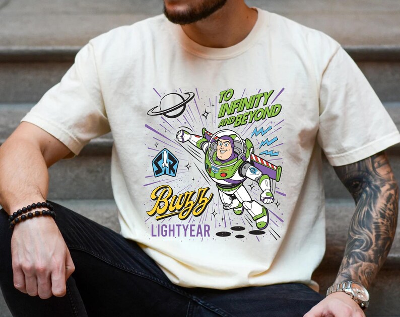 May include: A cream-colored t-shirt featuring a graphic of Buzz Lightyear in a flying pose. The design includes the text "TO INFINITY AND BEYOND" in green, "Buzz" in yellow script, and "LIGHTYEAR" in purple, along with a planet and star icons.
