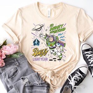 May include: A cream-colored t-shirt featuring a graphic of Buzz Lightyear in flight. The design includes the text "TO INFINITY AND BEYOND" and "BUZZ LIGHTYEAR," along with a planet, stars, and a space ranger emblem in shades of green, purple, and blue.