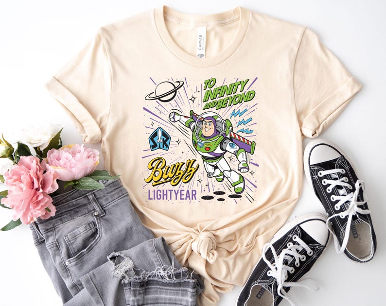 May include: A cream-colored t-shirt featuring a graphic of Buzz Lightyear in flight. The design includes the text "TO INFINITY AND BEYOND" and "BUZZ LIGHTYEAR," along with a planet, stars, and a space ranger emblem in shades of green, purple, and blue.