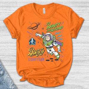 May include: An orange short-sleeve t-shirt featuring a graphic of Buzz Lightyear in flight. The design includes the text "TO INFINITY AND BEYOND", "Buzz", and "LIGHTYEAR", along with a planet icon and a space ranger emblem in blue, green, and purple.