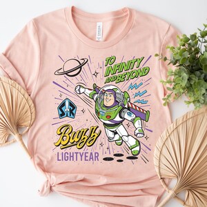 May include: A light peach-colored t-shirt featuring a graphic of Buzz Lightyear in a space suit. The design includes the text "TO INFINITY AND BEYOND" in green, "BUZZ" in yellow, and "LIGHTYEAR" in purple, along with a planet and rocket icon.