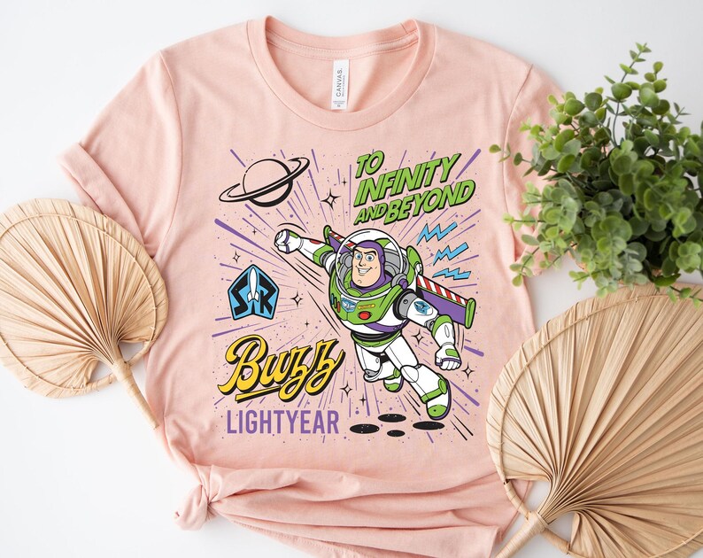 May include: A light peach-colored t-shirt featuring a graphic of Buzz Lightyear in a space suit. The design includes the text "TO INFINITY AND BEYOND" in green, "BUZZ" in yellow, and "LIGHTYEAR" in purple, along with a planet and rocket icon.