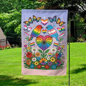 May include: A garden flag featuring a vibrant design with rainbow-colored birds perched above a heart-shaped flower. Butterflies and flowers in various colors surround the birds, with Pride flags and symbols throughout. The flag is displayed outdoors.