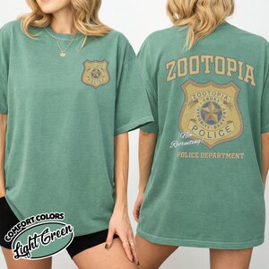 Puede incluir: Camiseta verde claro con una insignia de la polic&iacute;a de Zootopia en la parte delantera y trasera. La parte trasera de la camiseta muestra la palabra "ZOOTOPIA" encima de un dise&ntilde;o de insignia m&aacute;s grande con las palabras "TRUST, INTEGRITY, BRAVERY, POLICE, y POLICE DEPARTMENT."