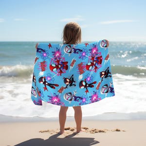 Puede incluir: Una toalla de playa rectangular de color azul claro con un patr&oacute;n repetitivo de personajes de dibujos animados con tem&aacute;tica de ara&ntilde;a en rojo, negro y blanco, intercalados con telara&ntilde;as rosas y la palabra "DAVIL" en una fuente roja y negrita.