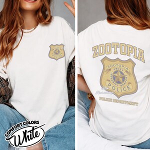 Puede incluir: Camiseta blanca con una insignia del Departamento de Polic&iacute;a de Zootopia en la parte delantera y trasera. La insignia delantera es m&aacute;s peque&ntilde;a, mientras que la trasera muestra el logotipo de Zootopia y las palabras "Integridad, Valent&iacute;a, Confianza". La camiseta tiene las palabras "Comfort Colors" y "White".
