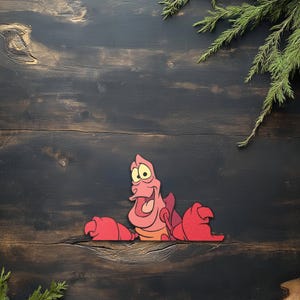 May include: A cartoon cutout of a red crab with a smiling expression, yellow eyes, and open mouth. The crab has large red claws and is set against a dark wood background with green sprigs.