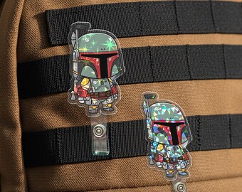 May include: Two cartoon-style badge reels featuring a character in a helmet and armor. The character has a red T-shaped visor and is set against a brown and black background. The badge reels are attached to a brown backpack.