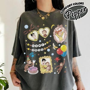 May include: Dark gray t-shirt with a colorful graphic featuring cartoon characters, balloons, and the text "name 2025". The shirt has a relaxed fit and the words "Comfort Colors Pepper" in a circular design.