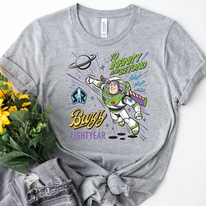 May include: A heather gray t-shirt featuring a graphic of Buzz Lightyear in a flying pose. The design includes the text "TO INFINITY AND BEYOND" in green, "Buzz" in yellow, and "LIGHTYEAR" in purple, along with a planet and starburst accents.