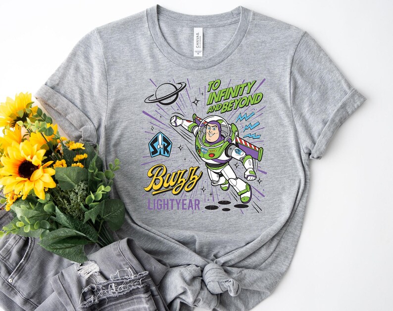 May include: A heather gray t-shirt featuring a graphic of Buzz Lightyear in a flying pose. The design includes the text "TO INFINITY AND BEYOND" in green, "Buzz" in yellow, and "LIGHTYEAR" in purple, along with a planet and starburst accents.