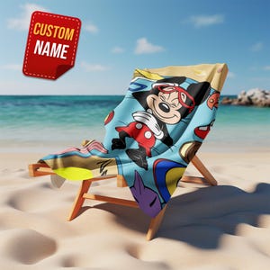 May include: A beach towel draped over a wooden lounge chair featuring a cartoon mouse wearing red goggles and red shorts. The towel has a light blue background with various colorful shapes and patterns. A red tag in the corner displays the text CUSTOM NAME.