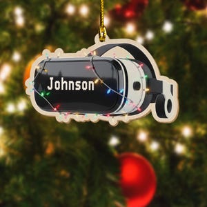 May include: A wooden VR headset Christmas ornament with the name "Johnson" in white. The black and white headset is decorated with colorful string lights. The ornament hangs from a gold string against a blurred Christmas tree background.