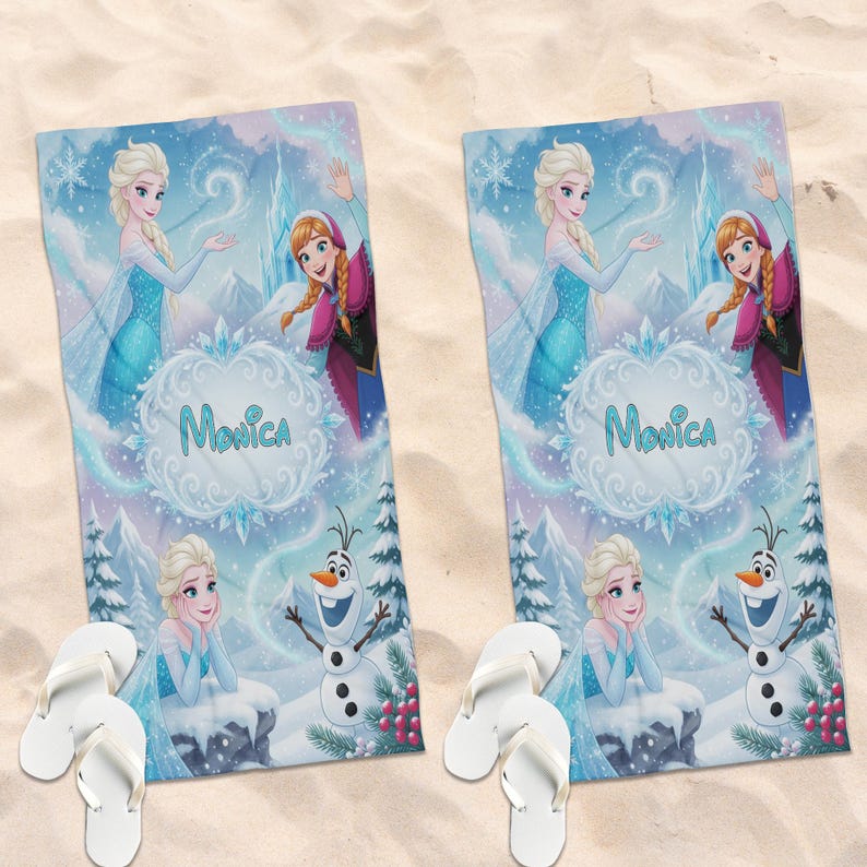 May include: Two beach towels featuring Elsa, Anna, and Olaf from Frozen, with the name "Monica" in a decorative font. The towels have a light blue and purple winter scene design. White flip-flops are placed near the towels on a sandy surface.