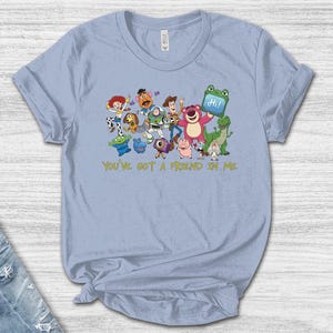 May include: A light blue t-shirt featuring a colorful graphic of various Toy Story characters, including Woody, Buzz Lightyear, and Lotso, with the text "YOU'VE GOT A FRIEND IN ME" printed in yellow below the characters and a blue tablet displaying "Hi!"
