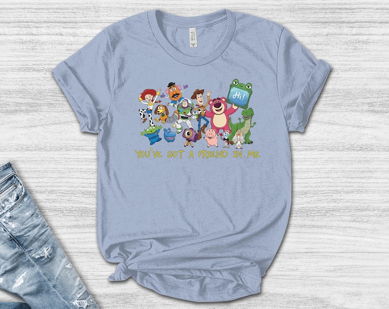 May include: A light blue t-shirt featuring a colorful graphic of various Toy Story characters, including Woody, Buzz Lightyear, and Lotso, with the text "YOU'VE GOT A FRIEND IN ME" printed in yellow below the characters and a blue tablet displaying "Hi!"