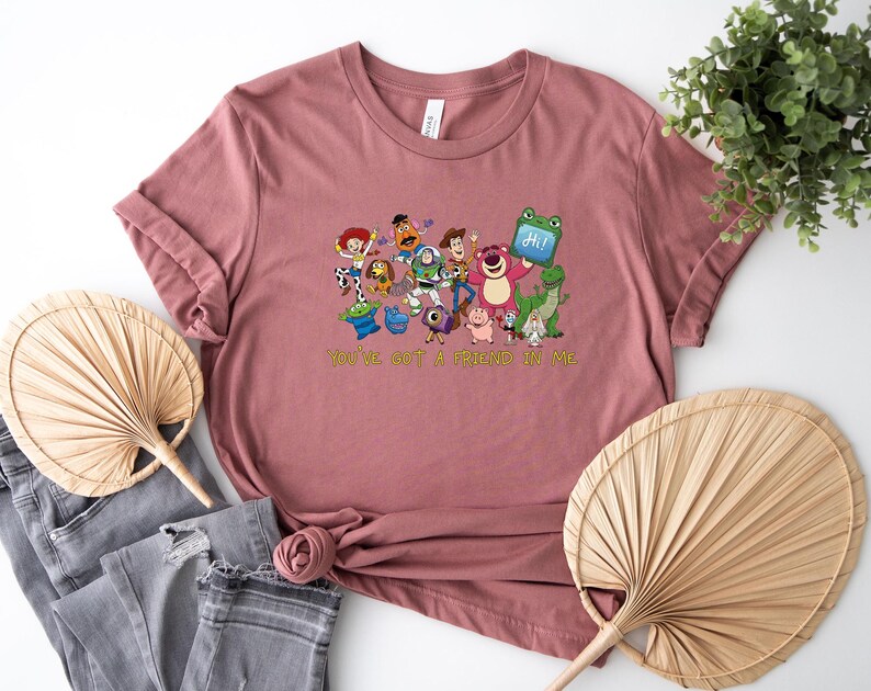 May include: A dusty rose short-sleeved t-shirt featuring a colorful graphic print of various animated toy characters and the text "YOU'VE GOT A FRIEND IN ME" in yellow lettering. The shirt has a crew neckline and a casual, relaxed fit design.
