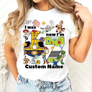 May include: White t-shirt featuring a colorful graphic of Toy Story characters and the text "I was now I'm" with the number 7. The design includes Woody, Buzz Lightyear, and other characters from the movie.