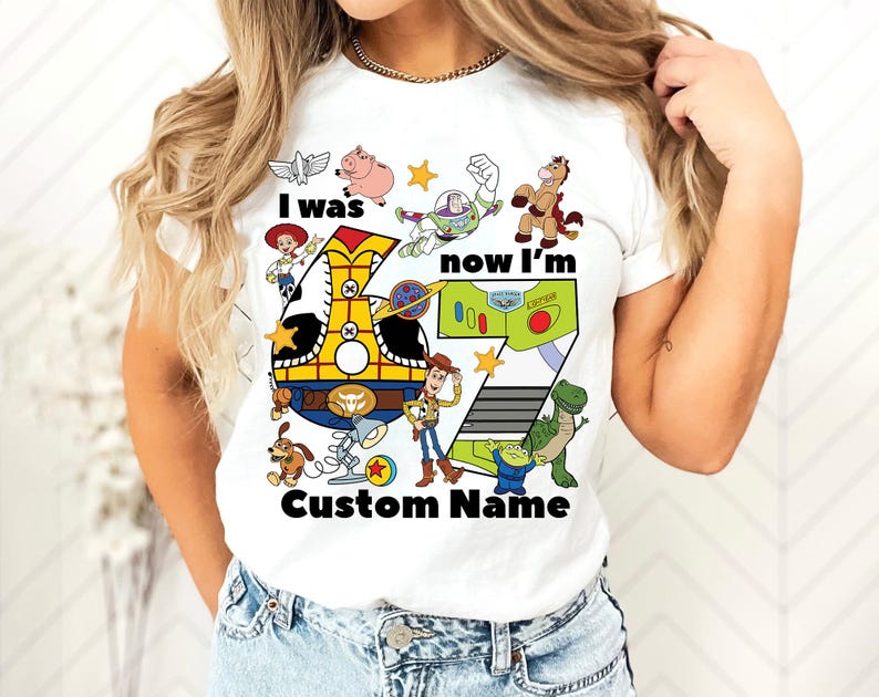 May include: White t-shirt featuring a colorful graphic of Toy Story characters and the text "I was now I'm" with the number 7. The design includes Woody, Buzz Lightyear, and other characters from the movie.