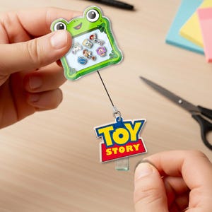 May include: A green frog-shaped badge holder with a retractable cord, holding several small character charms. The badge holder is connected to a "Toy Story" logo charm. The background includes a wooden surface, scissors, and sticky notes.