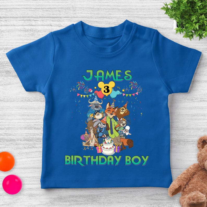 May include: A royal blue short-sleeve t-shirt featuring a graphic print of Zootopia characters wearing party hats. The text reads JAMES at the top, a yellow Mickey-shaped balloon with the number 3, and BIRTHDAY BOY at the bottom.