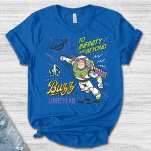 May include: A royal blue short-sleeve t-shirt featuring a graphic of Buzz Lightyear in a flying pose. The text reads "TO INFINITY AND BEYOND" in green, "Buzz" in yellow script, and "LIGHTYEAR" in purple. The design includes a planet and starbursts.