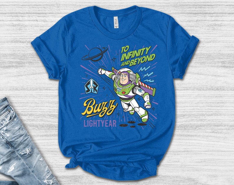 May include: A royal blue short-sleeve t-shirt featuring a graphic of Buzz Lightyear in a flying pose. The text reads "TO INFINITY AND BEYOND" in green, "Buzz" in yellow script, and "LIGHTYEAR" in purple. The design includes a planet and starbursts.