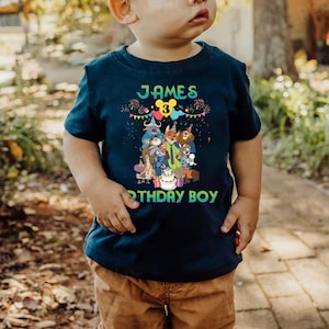 May include: A dark blue short-sleeved t-shirt featuring a colorful graphic of animated animal characters, a birthday cake, and the text "JAMES 3" and "RTHDAY BOY" in bright green letters, with a yellow Mickey Mouse ear icon above the number three.