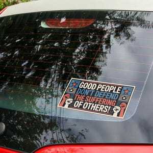 May include: A rectangular sticker with the text "GOOD PEOPLE DON'T DEFEND THE SUFFERING OF OTHERS!" in red, white, and blue. The sticker features stars, stripes, and raised fists. It is affixed to the rear window of a vehicle.