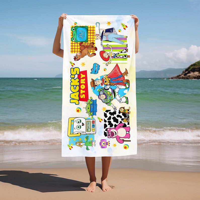 May include: White beach towel featuring colorful cartoon characters from "Toy Story." The towel has the text "Jack's Story" in yellow and red. The towel is being held up on a beach with the ocean in the background.