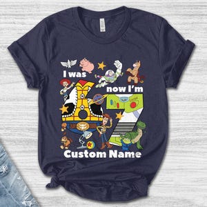 May include: Navy blue t-shirt featuring a colorful graphic design with characters and the text "I was now I'm 7 Custom Name". The design includes a train, a number 7, and various cartoon characters.
