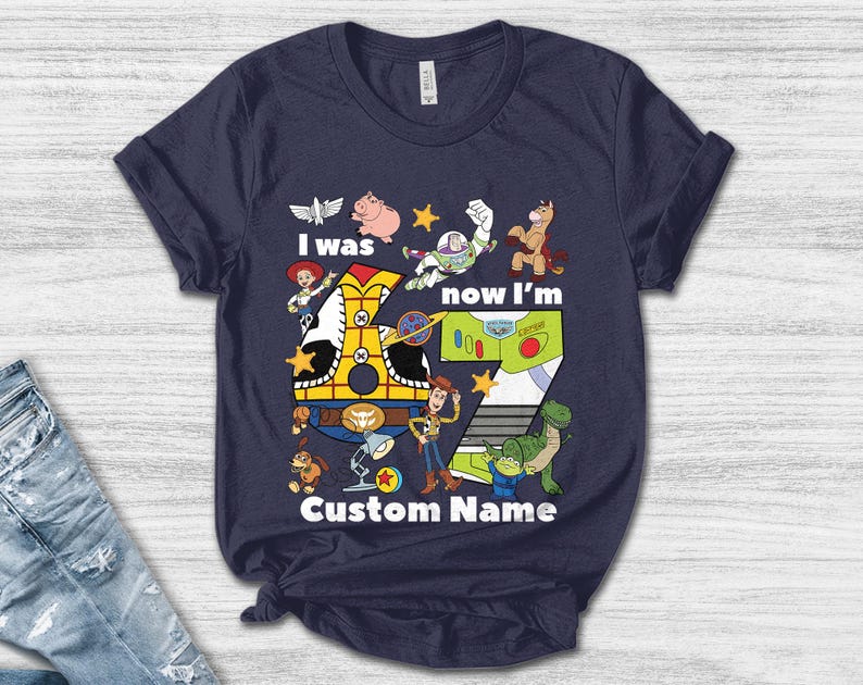 May include: Navy blue t-shirt featuring a colorful graphic design with characters and the text "I was now I'm 7 Custom Name". The design includes a train, a number 7, and various cartoon characters.