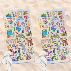 May include: Two white beach towels with a colorful Toy Story design, featuring characters like Buzz Lightyear and Woody, along with the word "JACK" repeated throughout. White flip-flops are at the bottom of each towel. The towels are on a sandy surface.