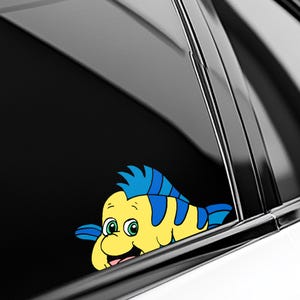 May include: A yellow cartoon fish sticker with blue accents, peeking out from behind a black surface. The fish has green eyes and a smiling expression. The sticker is likely for a car window or similar application.