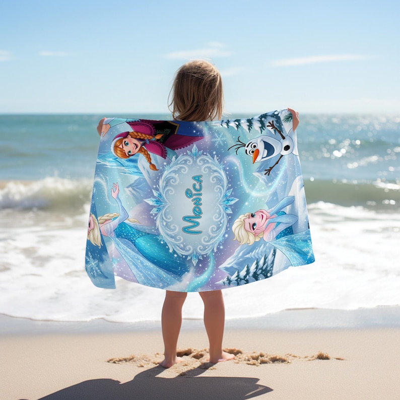 May include: A blue beach towel featuring characters from the movie Frozen, including Anna, Elsa, and Olaf. The towel has a personalized name, "Monica," in a decorative frame. The background shows a winter scene with snowflakes and mountains.