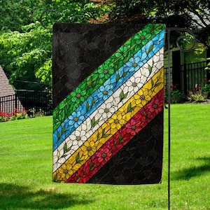 May include: A garden flag with a stained-glass design. The flag features diagonal stripes in green, blue, white, yellow, and red, each with floral patterns. The background is black with a textured appearance. The flag is displayed on a black metal stand.