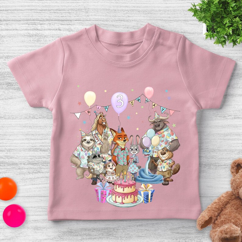 May include: Pink toddler t-shirt featuring a colorful graphic of cartoon animals celebrating a birthday. The design includes a cake, balloons, and the number 3. The shirt is made of soft material and has short sleeves.