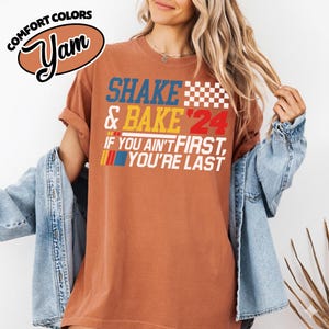 May include: Terracotta-colored t-shirt with the text "SHAKE & BAKE '24" in blue, yellow, and red. The shirt also features the phrase "IF YOU AIN'T FIRST, YOU'RE LAST" in white. The shirt has a vintage look with a comfort colors logo.