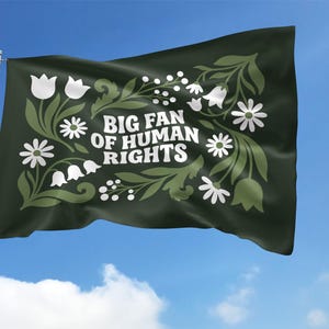 May include: A dark green flag waving in the wind against a blue sky. The flag features white floral designs and the text "BIG FAN OF HUMAN RIGHTS" in white. The flag is mounted on a silver pole.