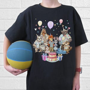 May include: Black t-shirt featuring a colorful cartoon illustration of various animal characters celebrating a birthday. The design includes a cake, balloons, and the number "3". The shirt is worn by a person holding a blue and yellow basketball.