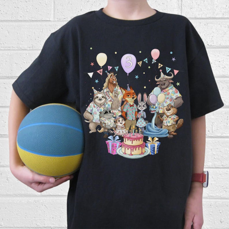 May include: Black t-shirt featuring a colorful cartoon illustration of various animal characters celebrating a birthday. The design includes a cake, balloons, and the number "3". The shirt is worn by a person holding a blue and yellow basketball.