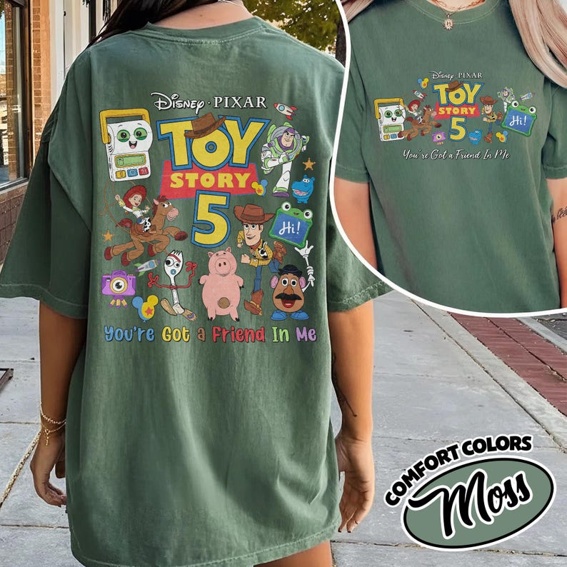 May include: A moss green t-shirt featuring a colorful graphic design from Disney Pixar's Toy Story 5. The design includes various characters and the text "You're Got a Friend In Me". The shirt is a Comfort Colors brand.