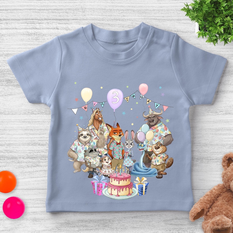 May include: A light blue toddler t-shirt featuring a colorful birthday party scene with cartoon animal characters. The number "3" is displayed on a balloon, and a birthday cake is also visible.